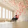 The Balloon Concerto Piano and its matching bench grace an elegant, sunlit room with large windows and a coffered ceiling. Pink balloons float mysteriously around the piano, evoking a whimsical and surreal atmosphere, as if readying for an enchanting performance.