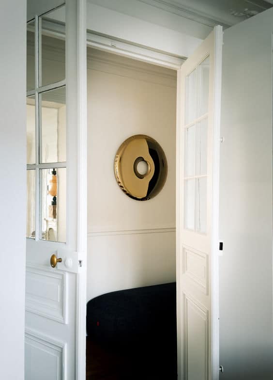 A minimalist white room featuring an open door with a glass pane, revealing a large, circular, Gold Donut Mirror inside.