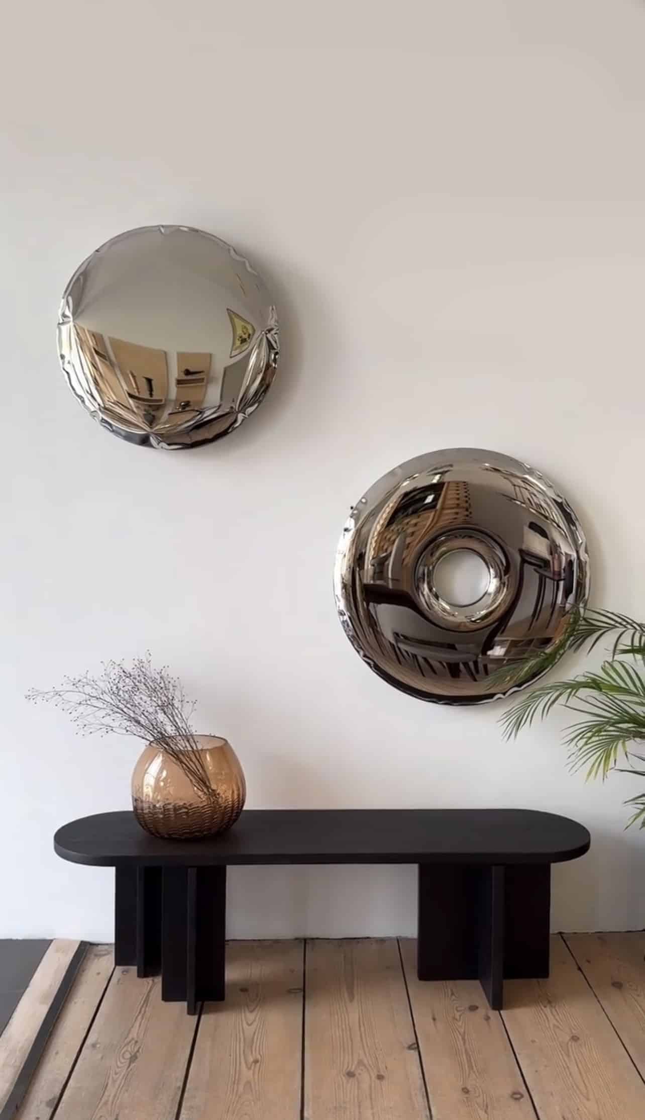 Two modern Solid Donut Mirrors above a black bench with a decorative vase and a plant on a wooden floor against a light grey wall.