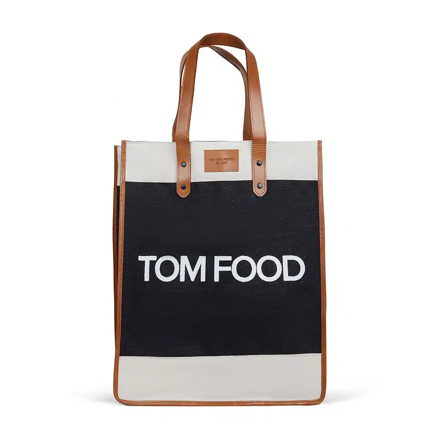 A rectangular Tom Food with brown leather handles and trim. The bag is predominantly black with a white canvas section at the back. The text "Tom Food" is printed in large white letters on the black front portion.