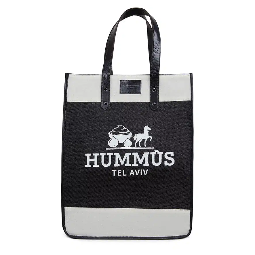 The "Hummus" tote bag is a black and white tote with black handles. It showcases a white graphic of a bowl of Mediterranean dip and a seated animal, accompanied by the text "HUMMUS TEL AVIV" printed in white.