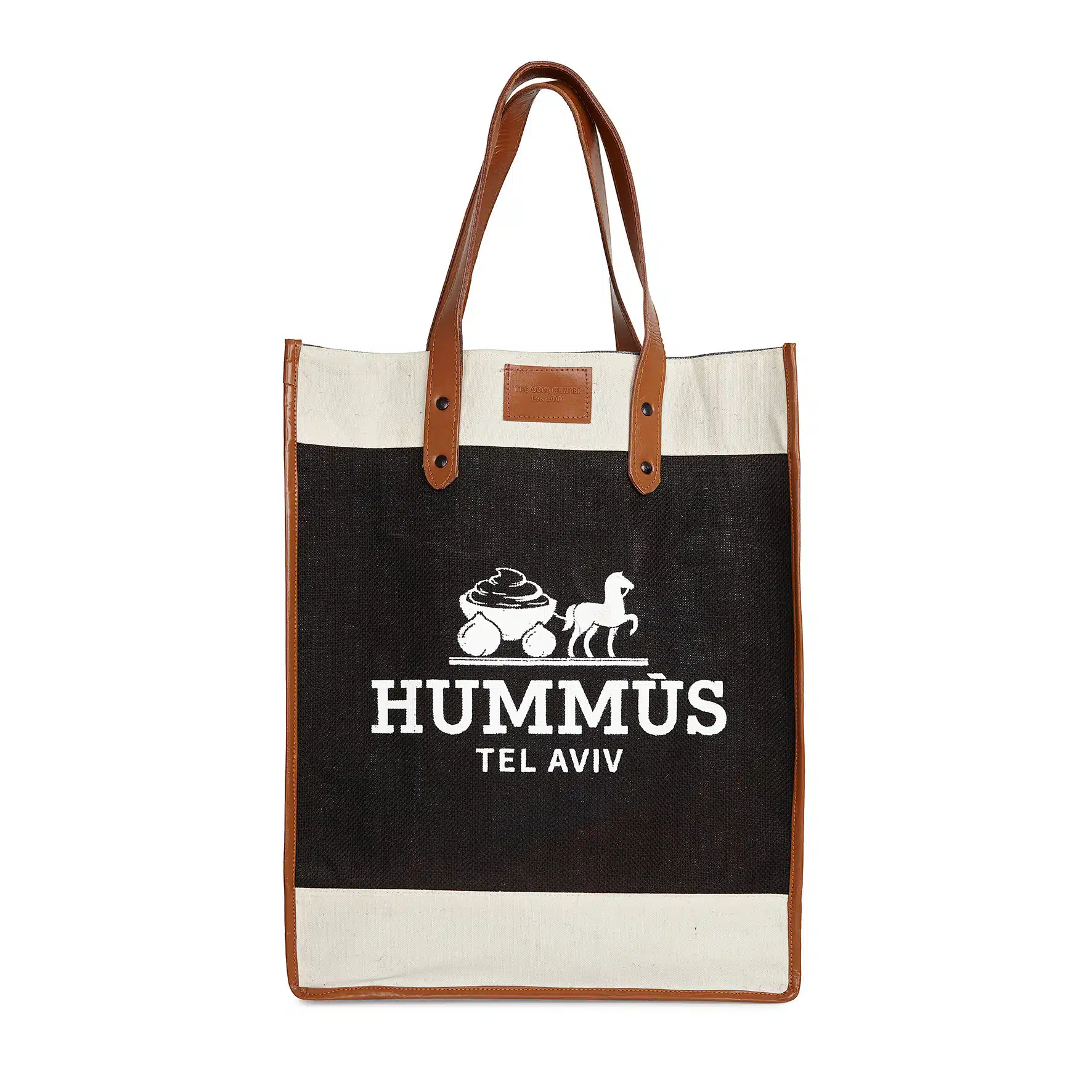 A black and white canvas tote bag with brown leather handles sits upright, featuring a graphic of a chickpea, tahini jar, and horse-drawn carriage above the bold white text "HUMMUS TEL AVIV," celebrating delicious Mediterranean food.