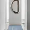 A modern, elongated Pebble Mirror 97 x 150 cm with a reflective metallic frame, mounted on a white wall at the end of a narrow hallway with a light blue runner rug on the floor.