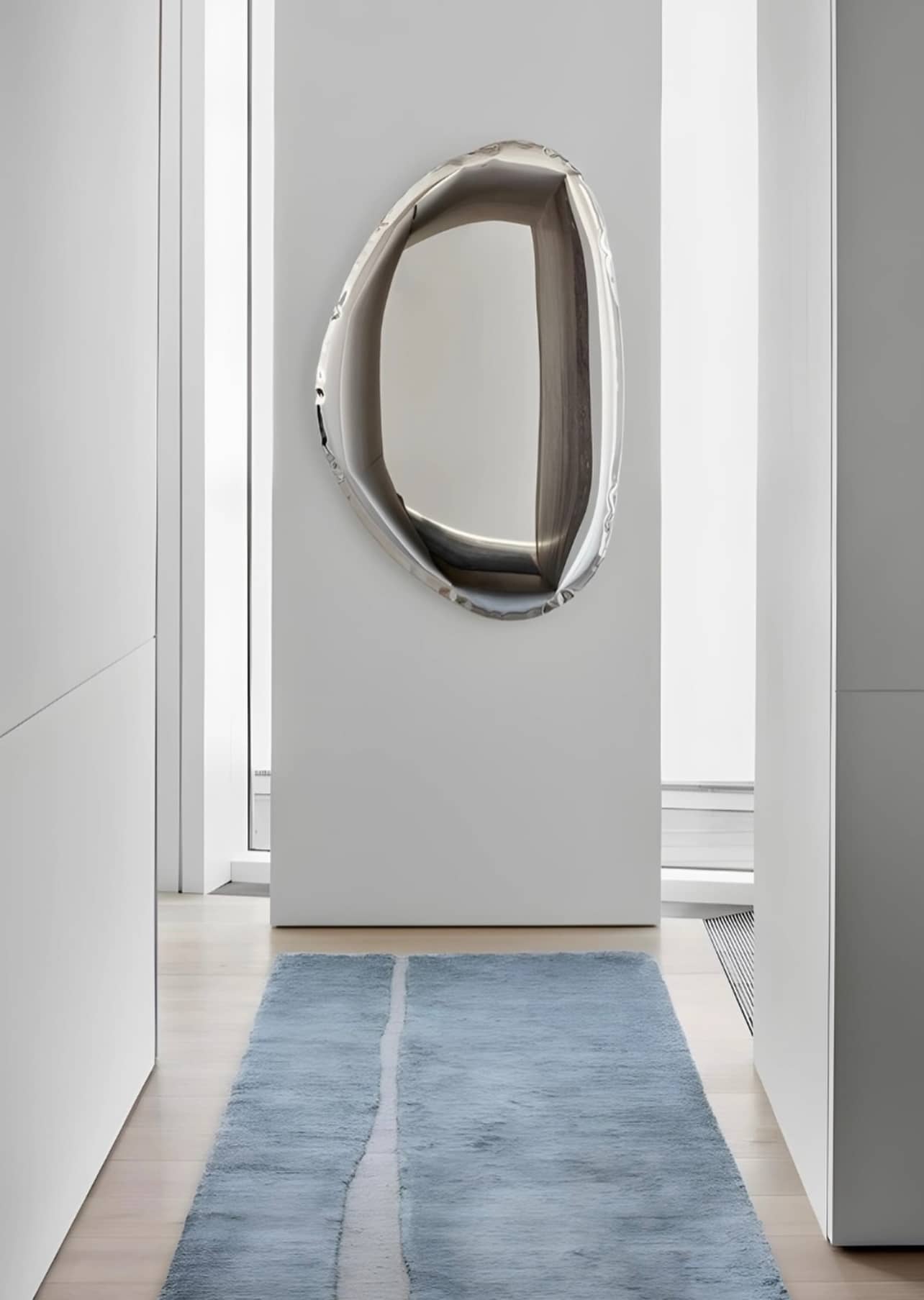 A modern, elongated Pebble Mirror 97 x 150 cm with a reflective metallic frame, mounted on a white wall at the end of a narrow hallway with a light blue runner rug on the floor.