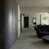 A minimalist living room with smooth, grey walls and floor, featuring dark wood cabinets, a curved black sofa, and a large Pebble Mirror 97 x 150 cm on the wall.