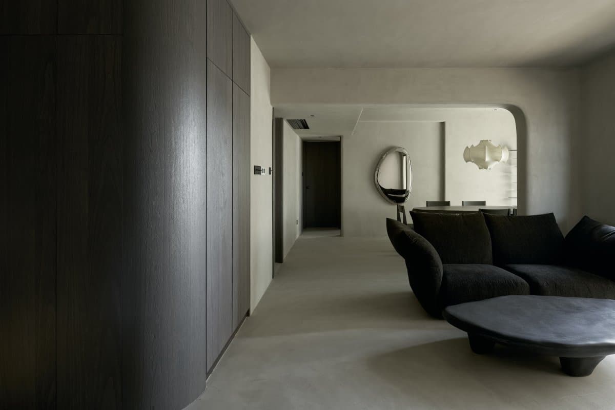 A minimalist living room with smooth, grey walls and floor, featuring dark wood cabinets, a curved black sofa, and a large Pebble Mirror 97 x 150 cm on the wall.