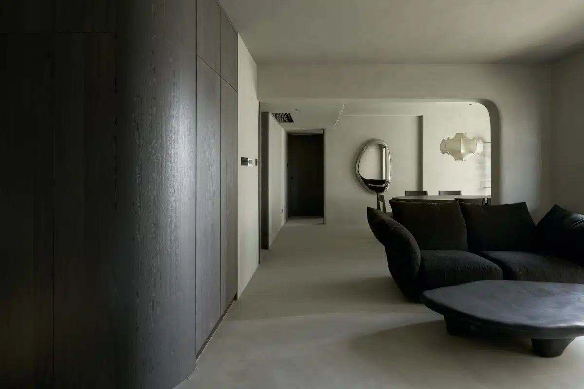 A minimalist living room with smooth, grey walls and floor, featuring dark wood cabinets, a curved black sofa, and a large Pebble Mirror 97 x 150 cm on the wall.