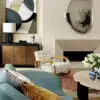 A modern living room features a unique teal sofa with mustard and gray pillows, a light wood coffee table with a vase of white tulips, and an abstract armchair beside a minimalist black and brown cabinet. A Pebble Mirror - 150 x 97 cm adorns the walls, completing the home decor.