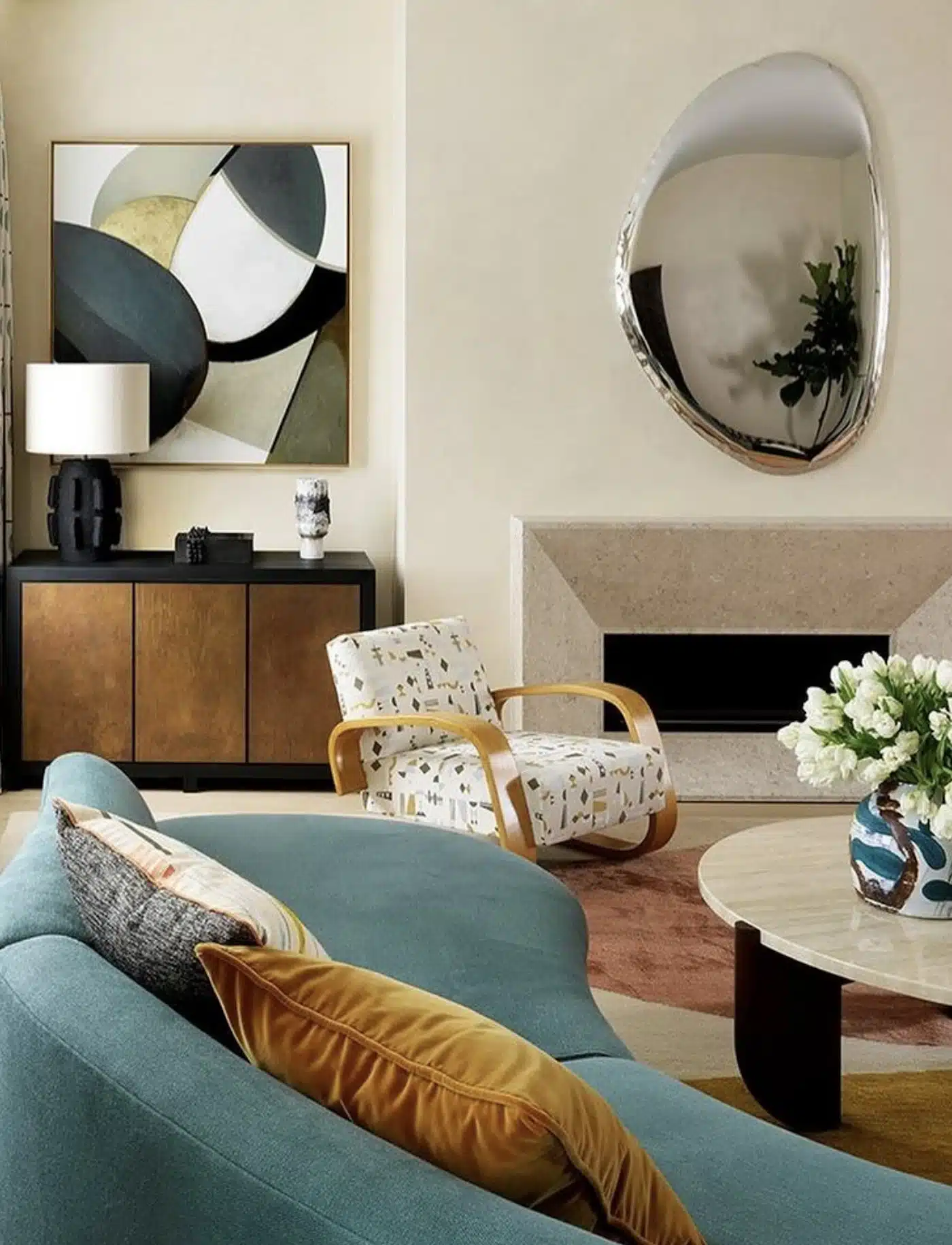 A modern living room features a unique teal sofa with mustard and gray pillows, a light wood coffee table with a vase of white tulips, and an abstract armchair beside a minimalist black and brown cabinet. A Pebble Mirror - 150 x 97 cm adorns the walls, completing the home decor.