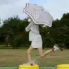 A person in a beige outfit leaps on yellow splash pads, holding a Karen Walker Umbrella over their head, creating a playful and surreal scene outdoors.