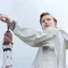 A person in a white jacket reaches up to adjust a Karen Walker Umbrella handle, set against a cloudy sky backdrop.