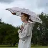 A woman wearing a beige raincoat holds a Karen Walker Super Duper Strength umbrella, intricately patterned. She stands outdoors with raindrops visible in the air, showcasing an attentive gaze to her side.