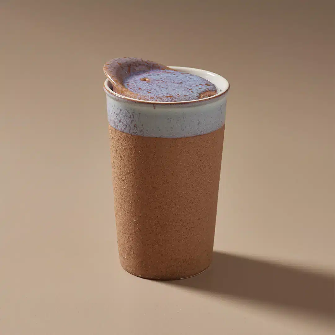 A Ceramic Keep Mug with a light blue lid and a brown body sits on a beige surface. The mug's simple design features a matte finish, and the lid appears to fit snugly on top, blending style with functionality. Shadows cast to the right suggest light from the left.