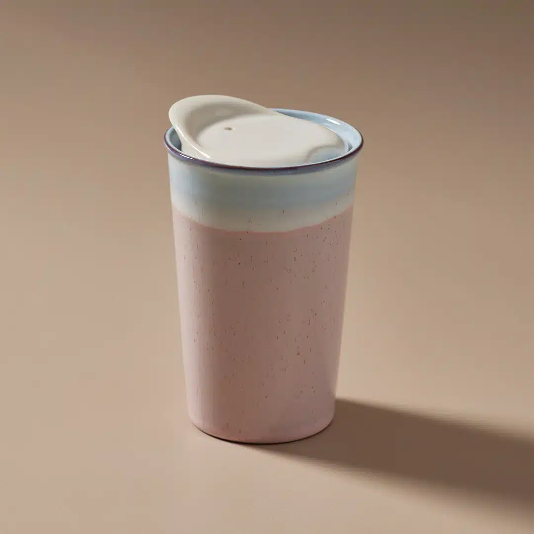 The Ceramic Keep Mug features a pastel pink and light blue gradient design, topped with a beige silicone lid. Placed against a neutral beige background, the mug casts a gentle shadow to the right, suggesting soft lighting from the left.