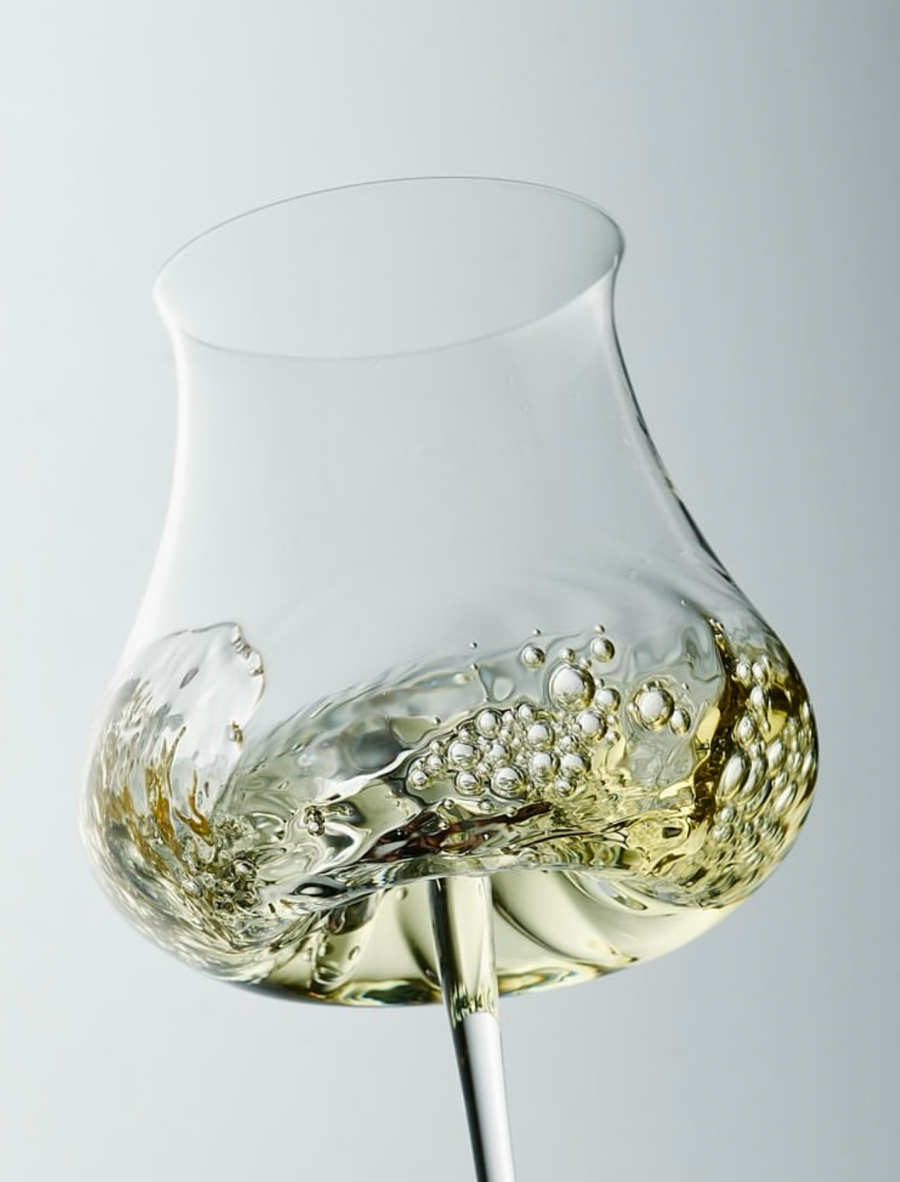 A clear wine glass with white wine swirls inside, creating bubbles and a dynamic pattern. The background is a soft, neutral gray, emphasizing the fluid motion of the liquid.